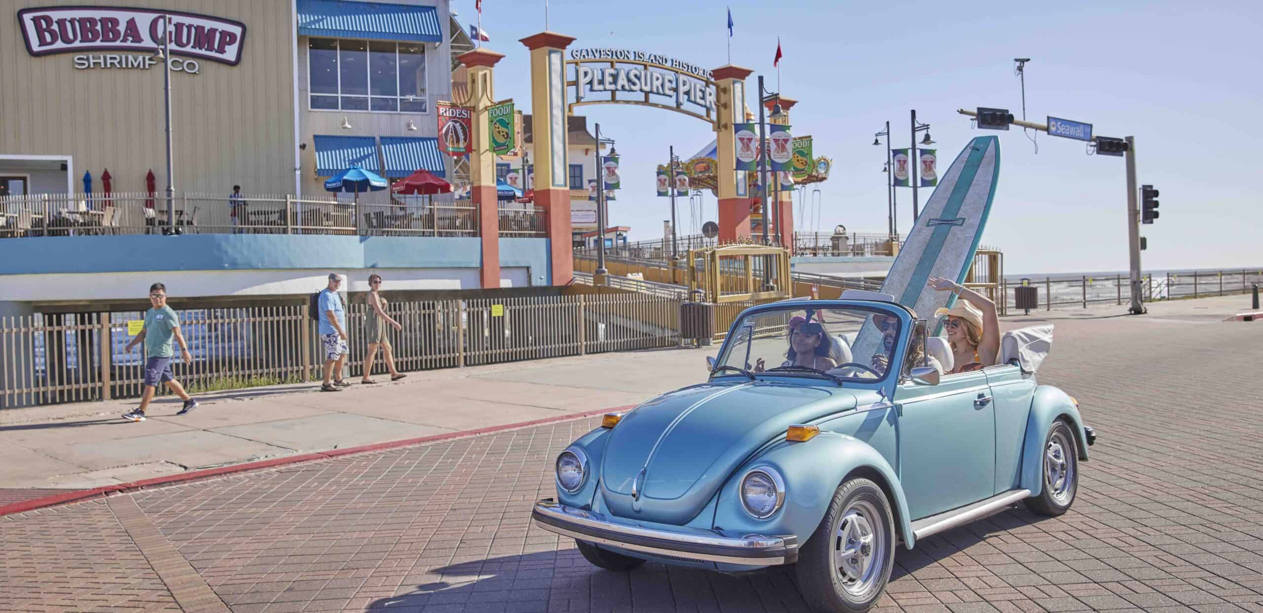 Blue vintage Volkswagen Beetle parked near the beach with amusement park in background.