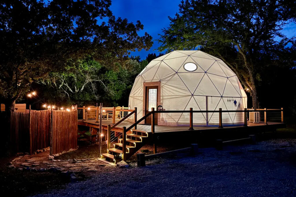 A lit wooden deck leads to a large geodesic dome tent at night, surrounded by trees and outdoor string lights.