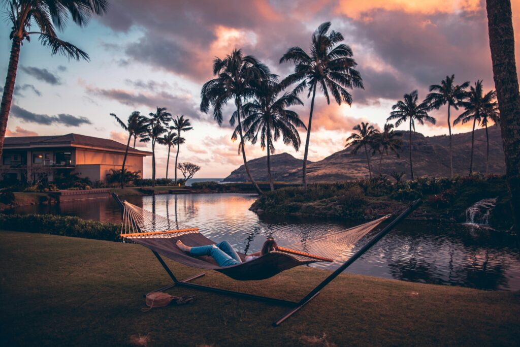 A person relaxes on a hammock beside a pond at sunset, surrounded by palm trees, with mountains and a building in the background.