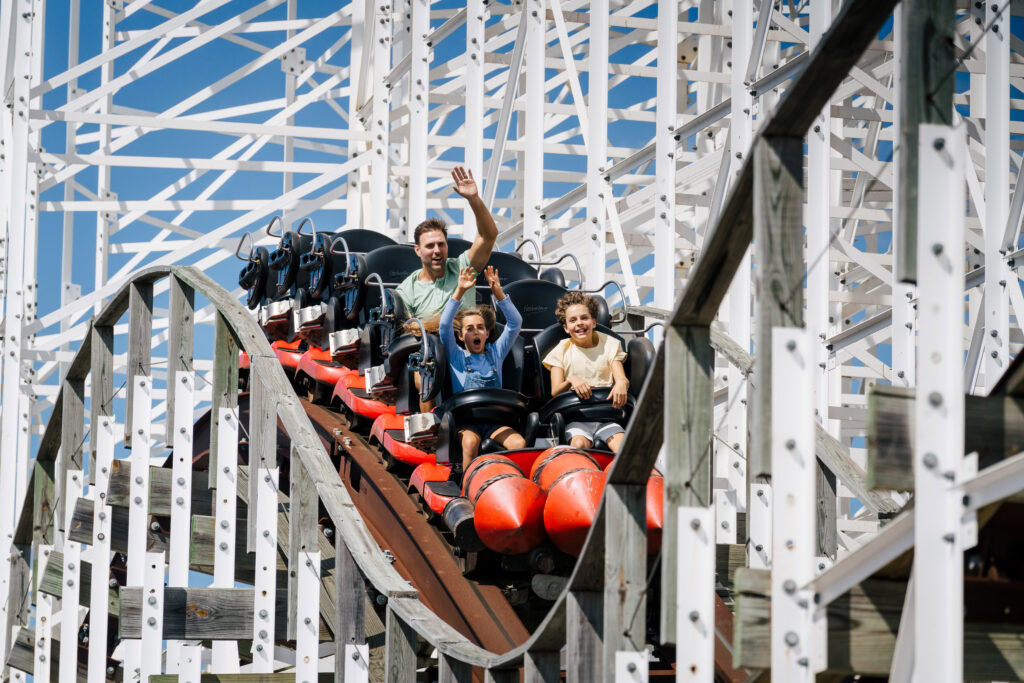 Three people ride a roller coaster downhill; one adult waves, and two children sit beside him. White wooden supports and blue sky are visible in the background.