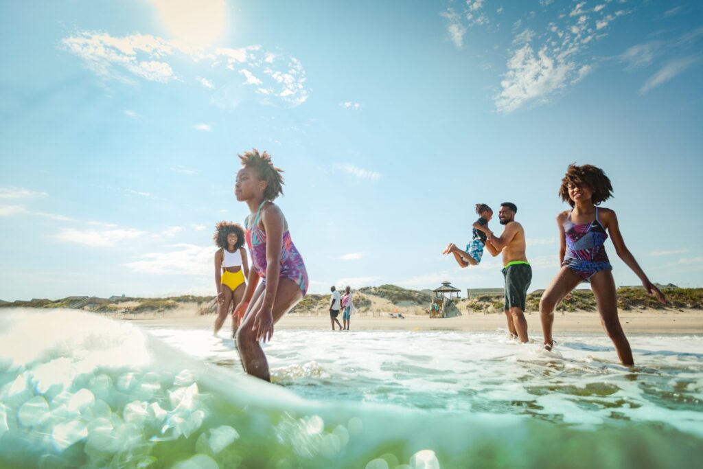 A group of people, including children and adults, stand and play in the shallow water at a sunny beach with blue sky and distant sand dunes.