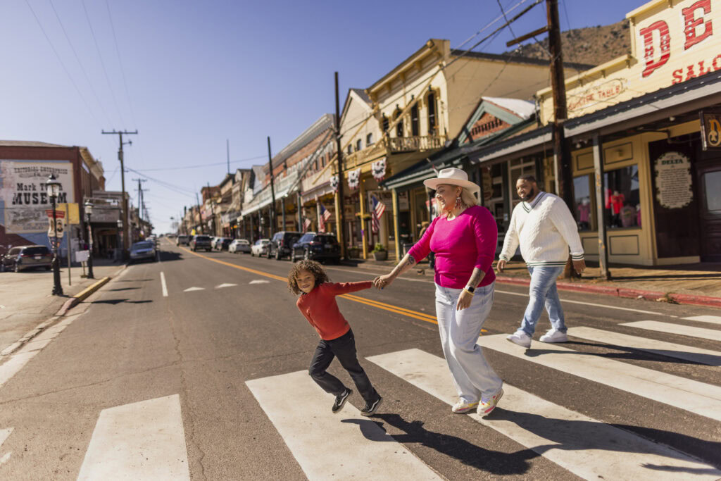 A woman in a cowboy hat and a child hold hands while crossing a street in a historic town, with another adult walking behind them.