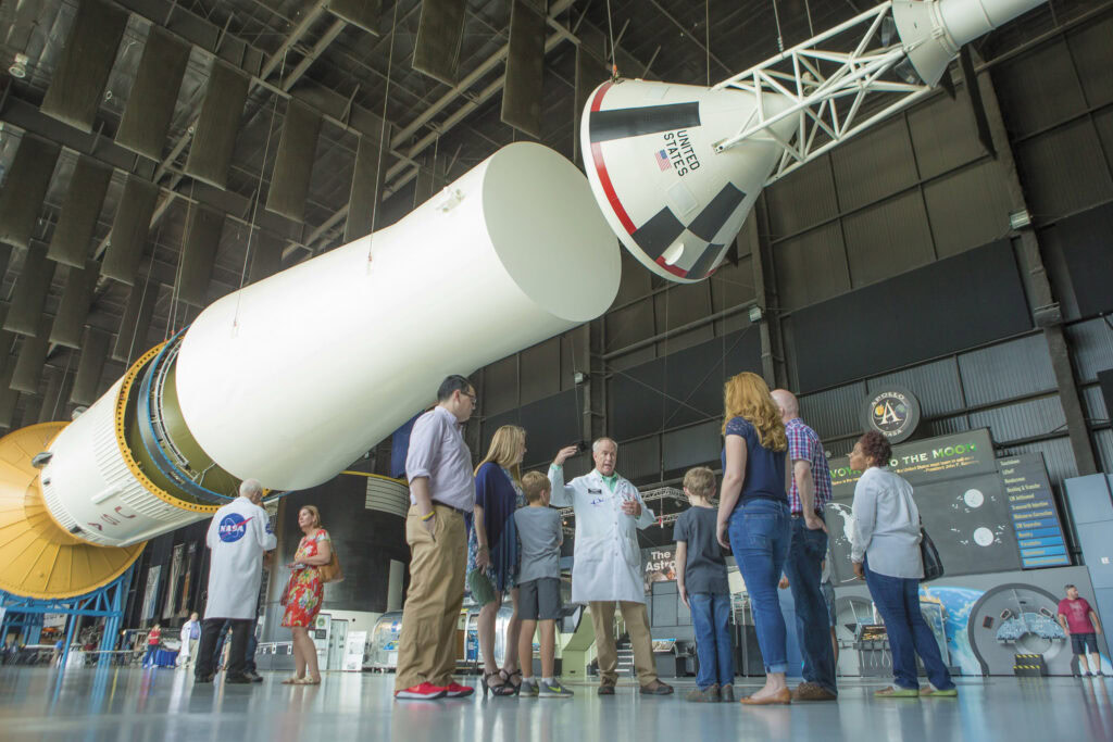 A group of people listen to a guide standing under a large suspended rocket exhibit inside a spacious museum hall.