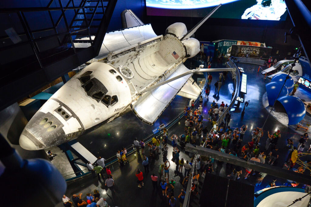 A crowd views the Space Shuttle Atlantis displayed indoors at a museum, with exhibits and informational signs visible in the background.