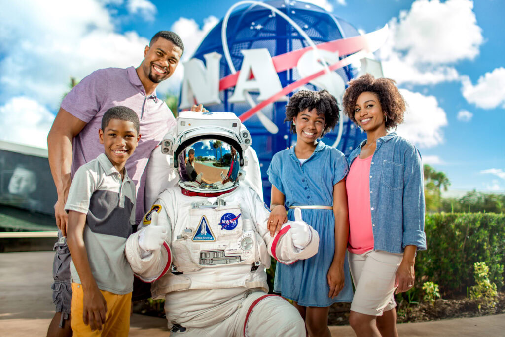 A family poses with a person in a NASA spacesuit in front of a NASA logo sign outdoors on a sunny day.