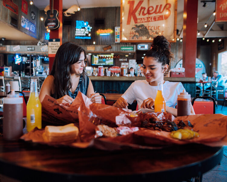 Two women sit at a table eating barbecue with bottled drinks in a casual restaurant decorated with neon signs and vintage memorabilia.