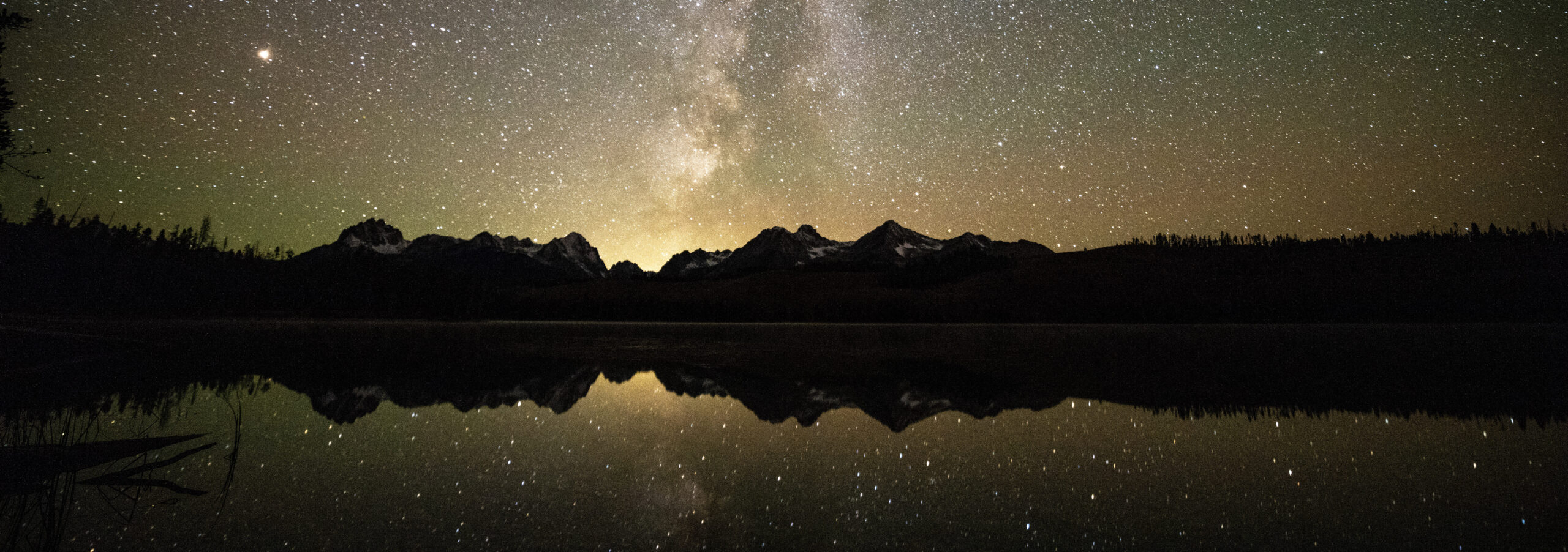 Starry night sky and Milky Way over mountain range, reflected in a calm lake, with silhouetted trees along the shore.