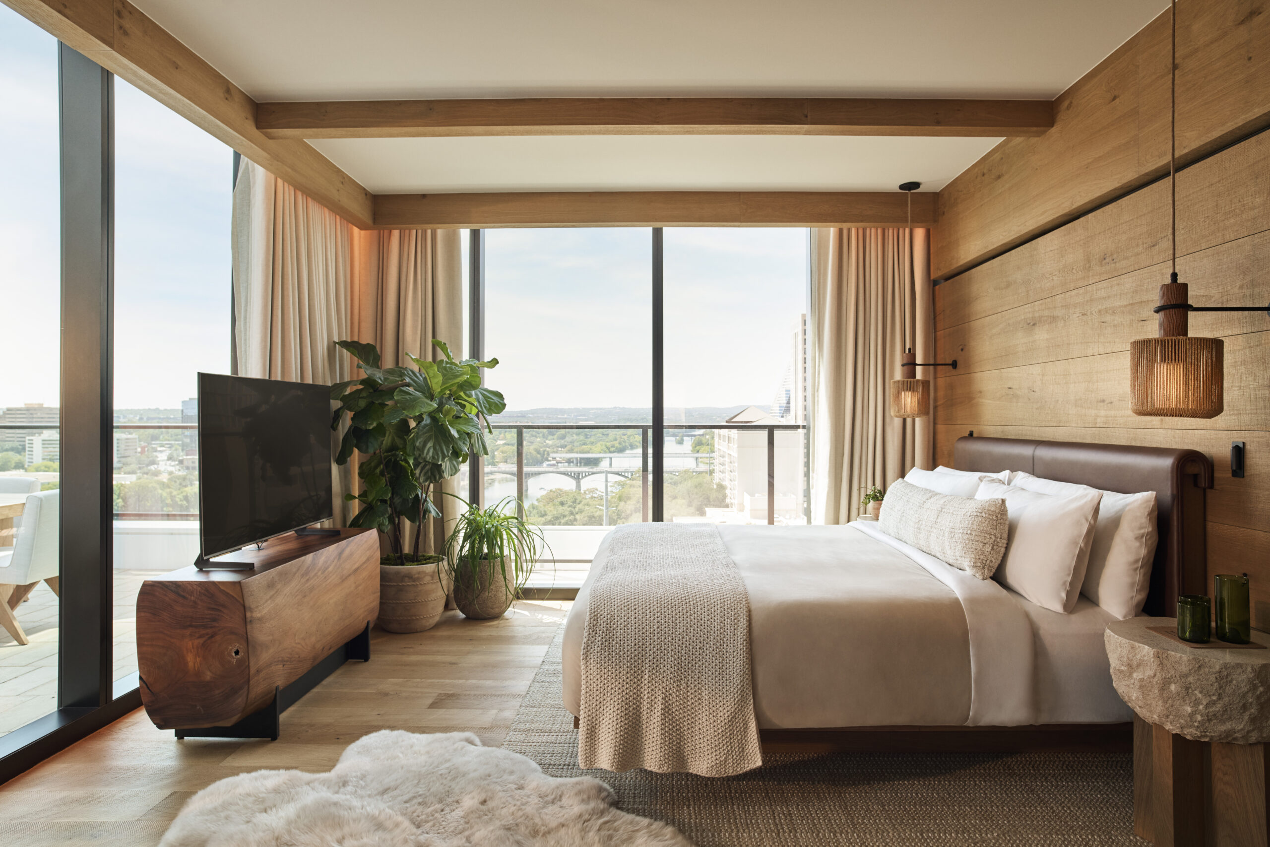 Modern bedroom with large windows, a king bed, wooden walls, a TV on a wooden stand, potted plants, and hanging lights. Balcony and city view are visible through the glass doors.