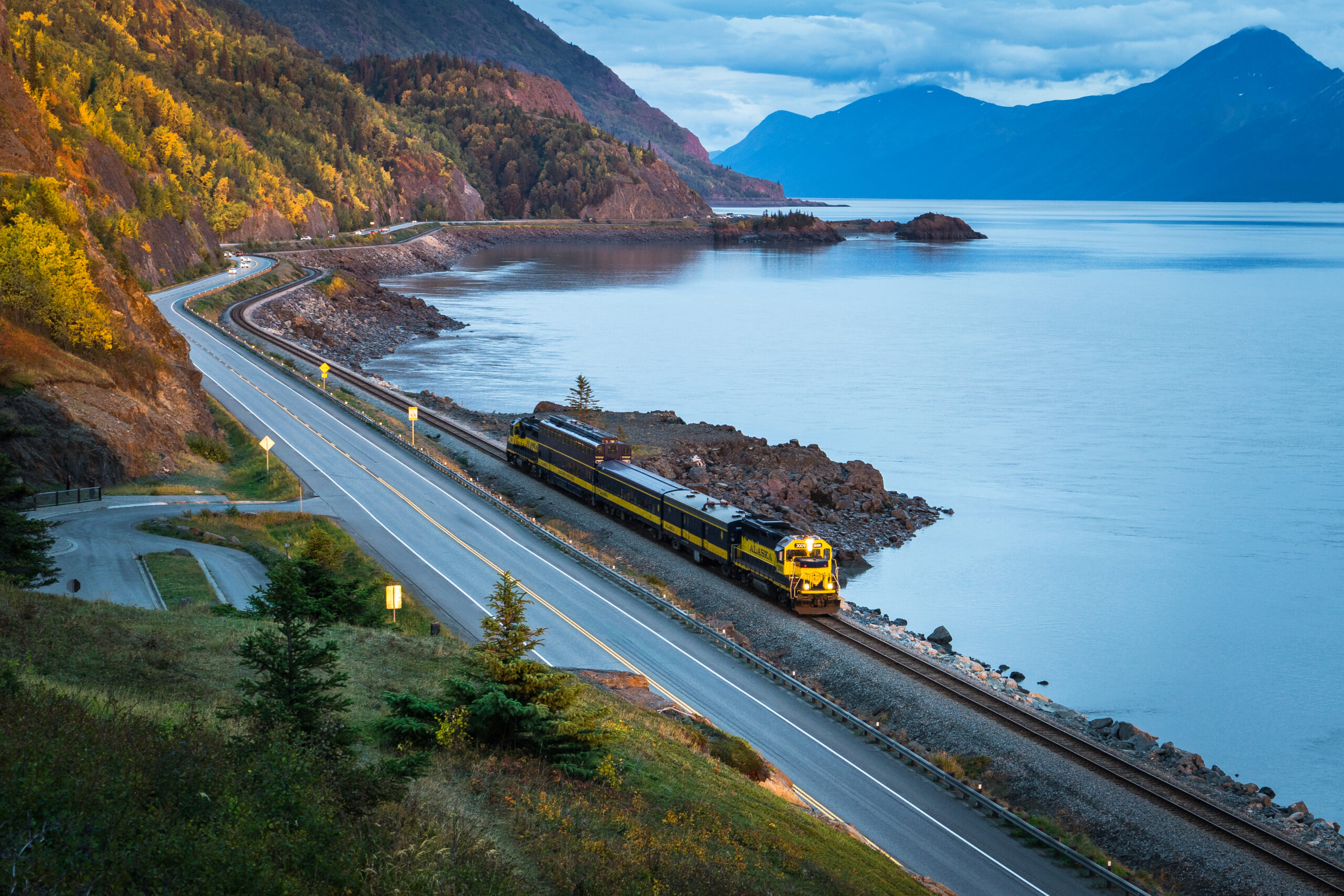 A yellow and blue train travels along tracks beside a winding road and a large lake, with mountains and trees in the background under a cloudy sky.