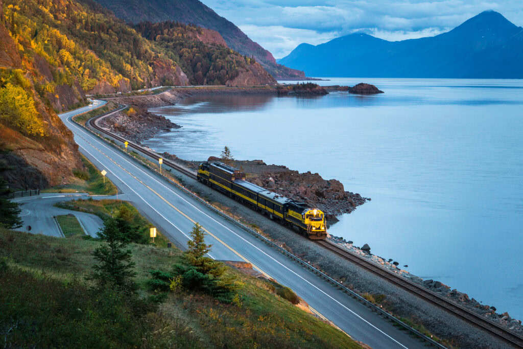 A yellow and blue train travels beside a winding road along a lake with mountains and autumn trees in the background under a cloudy sky.