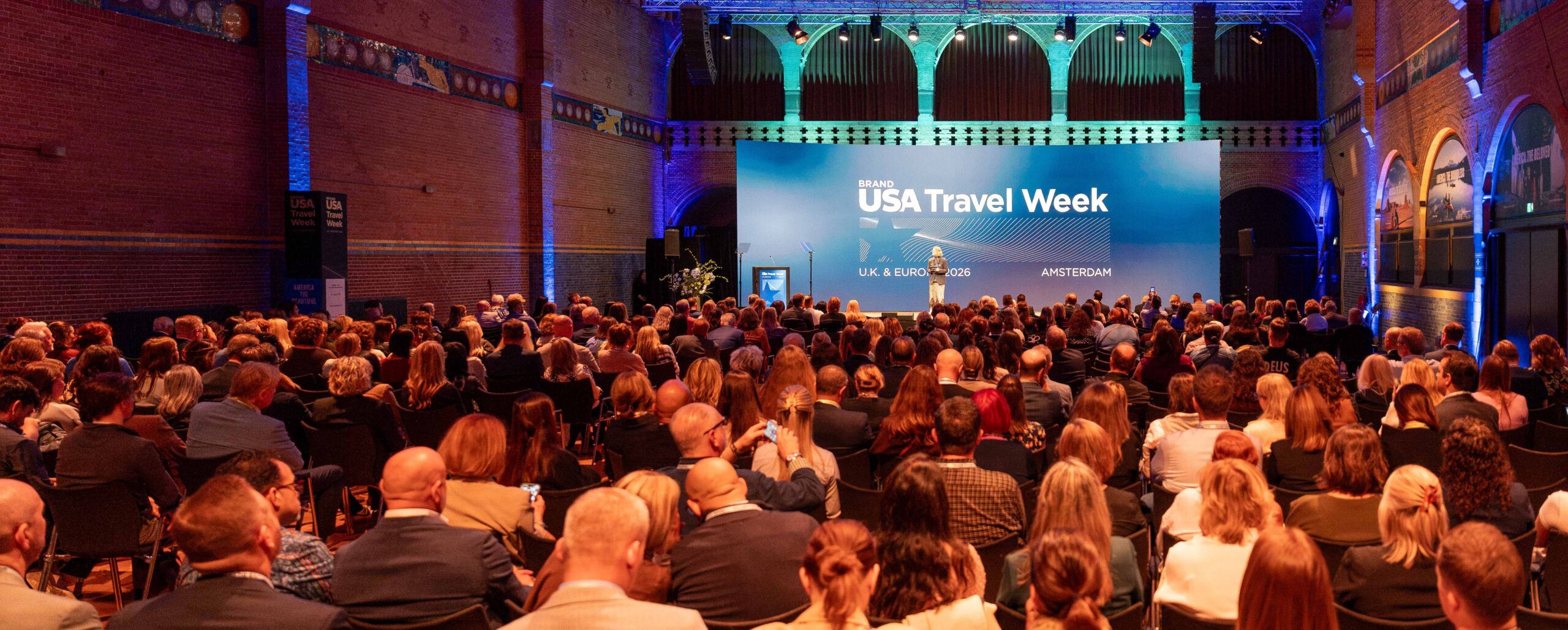 A large audience sits facing a stage with a blue backdrop reading “Brand USA Travel Week” in a spacious, well-lit venue.