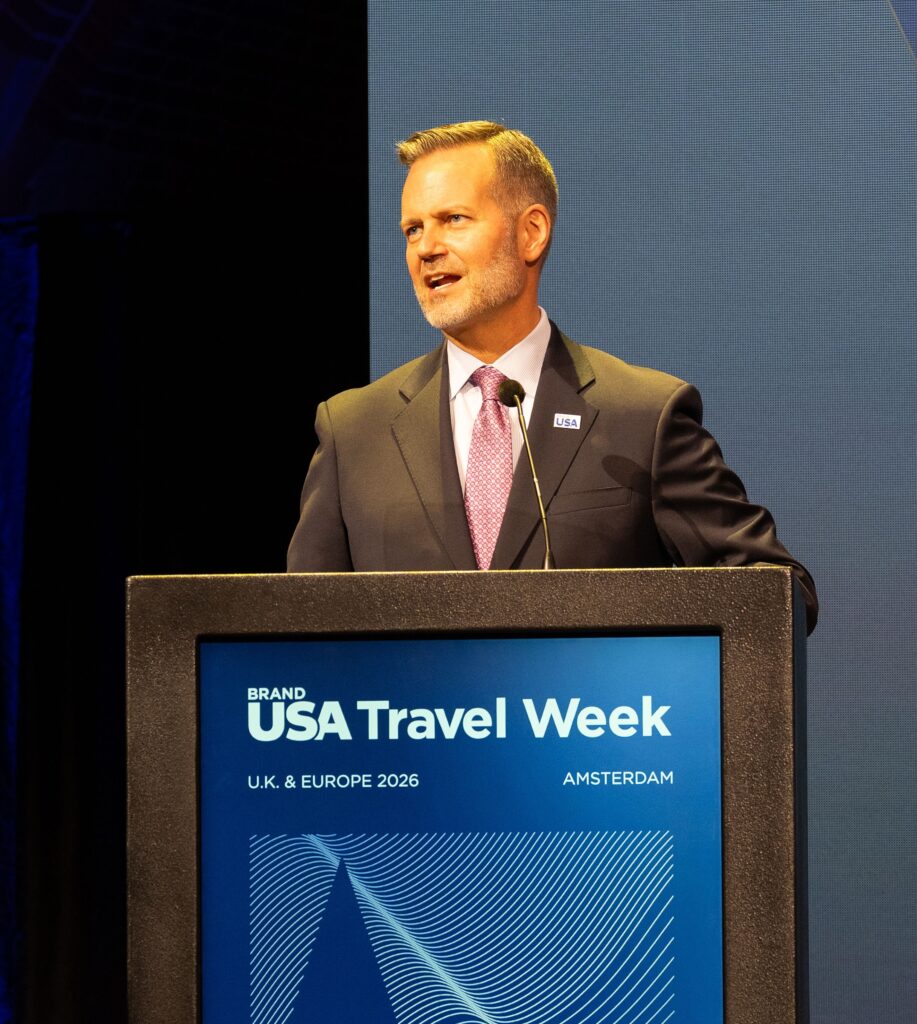 A man in a suit speaks at a podium with a sign reading “USA Travel Week, U.K. & Europe 2026, Amsterdam.”.