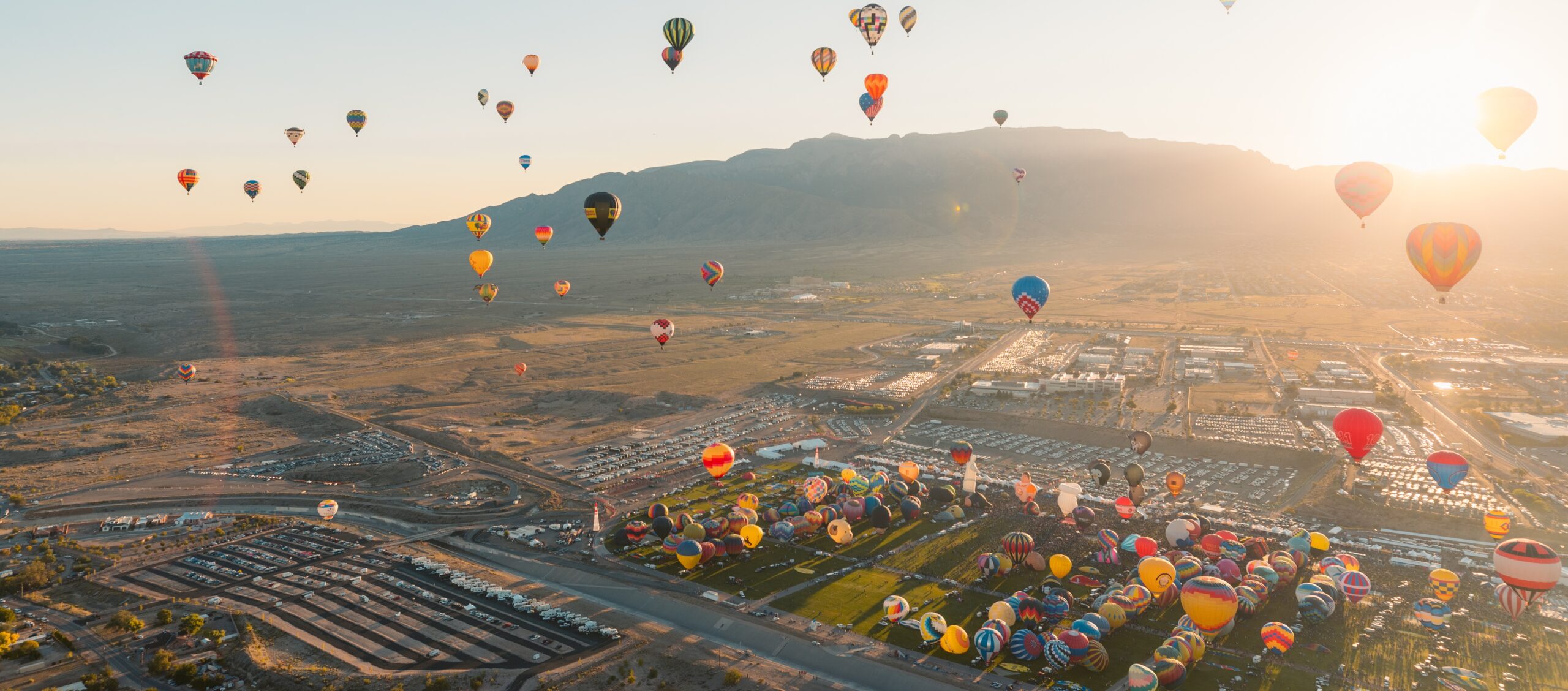 Dozens of colorful hot air balloons rise over a vast landscape at sunrise, with mountains and a city in the background.