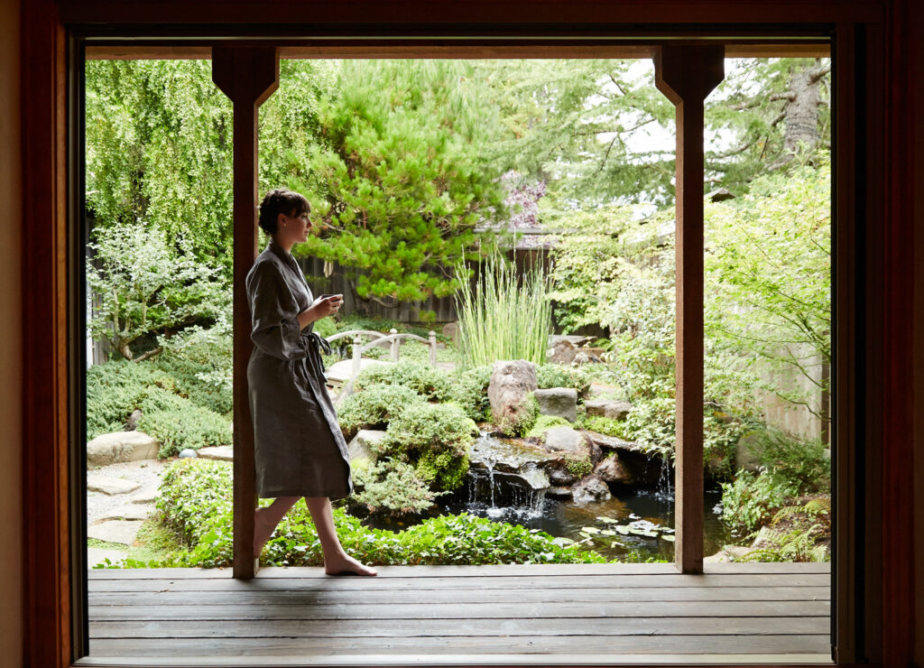A person in a robe holding a cup stands on a wooden porch, looking out at a lush garden with a small waterfall and pond.