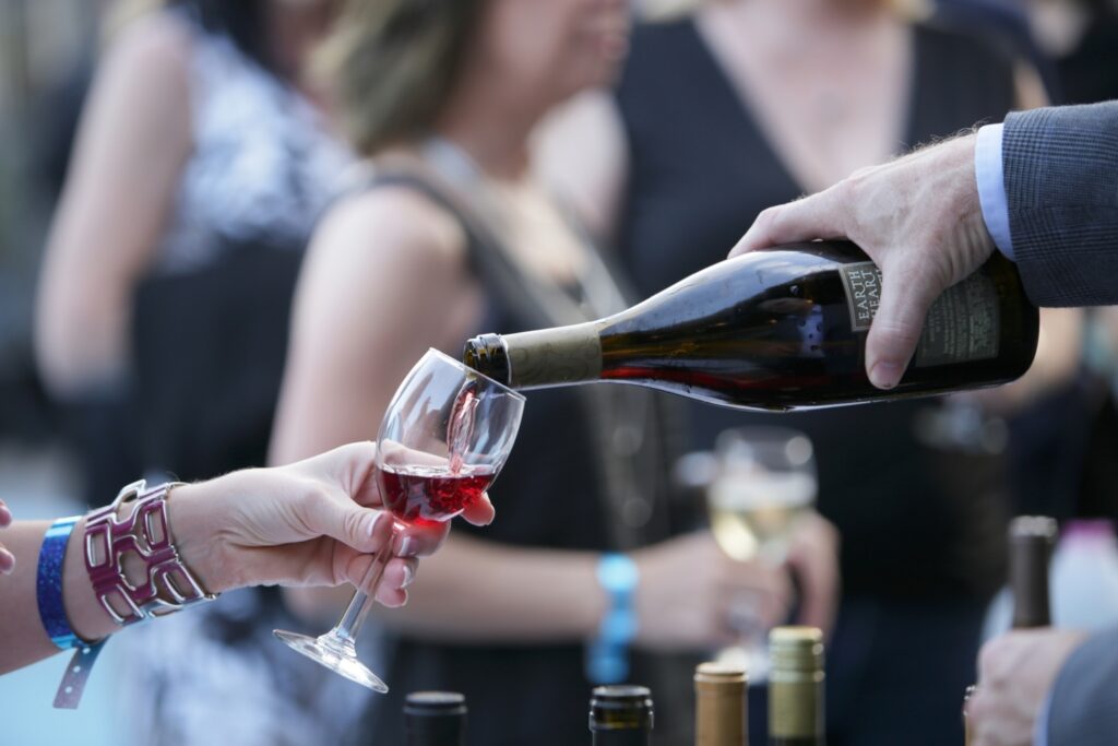 A person pours red wine into a glass held by another person at an outdoor social gathering, with more people and wine bottles in the background.