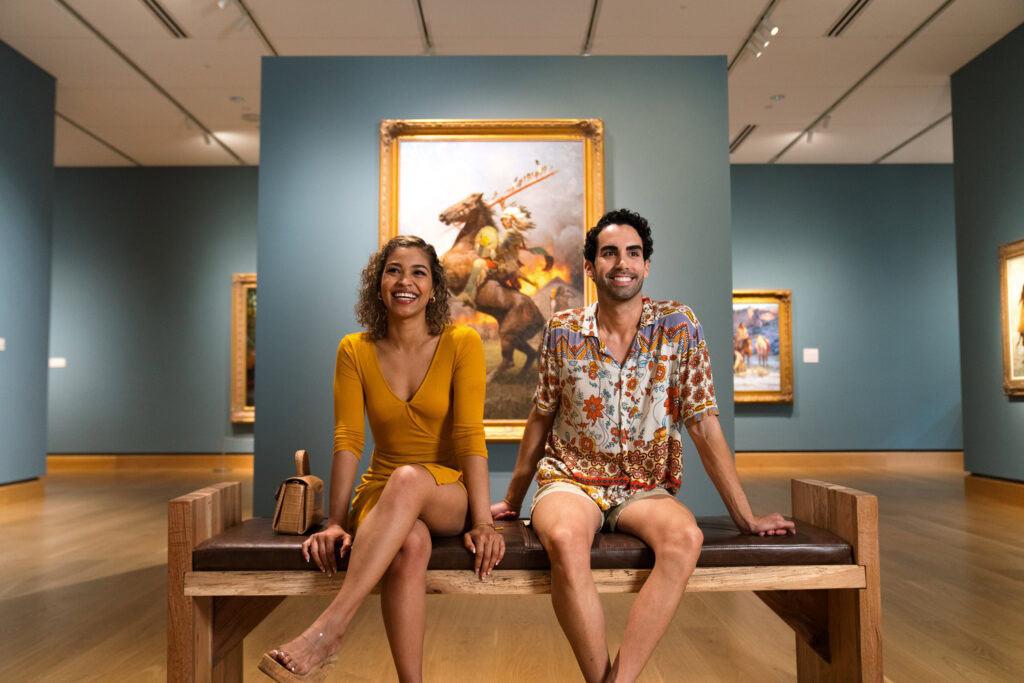 Two people sit on a wooden bench in an art gallery, smiling, with paintings in gold frames displayed on blue walls behind them.