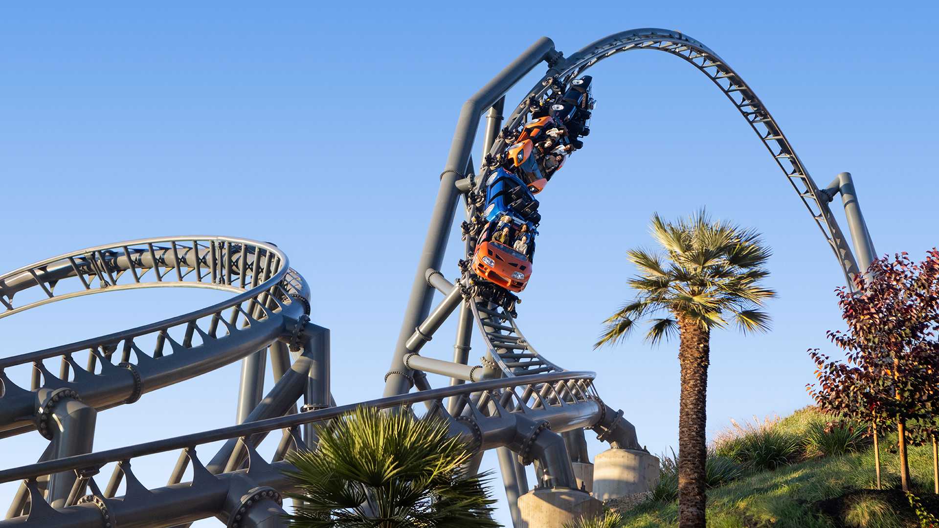 Roller coaster cars with riders navigate a steep, twisted loop on a steel track with palm trees and blue sky in the background.