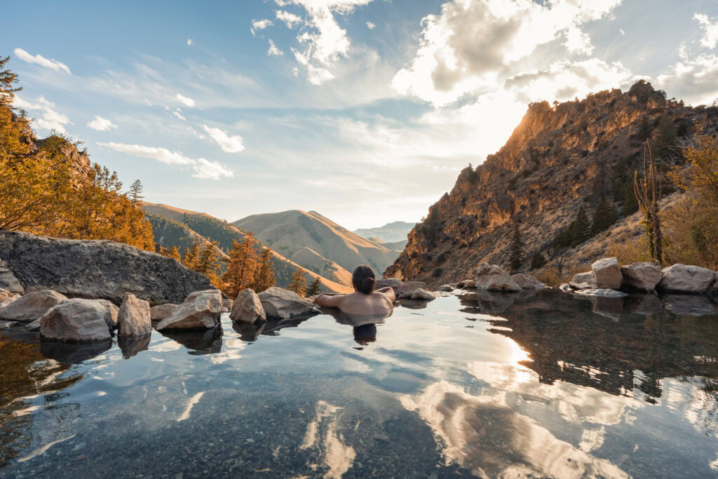 A person relaxes in a natural hot spring pool surrounded by rocks, overlooking mountainous terrain under a partly cloudy sky at sunset.