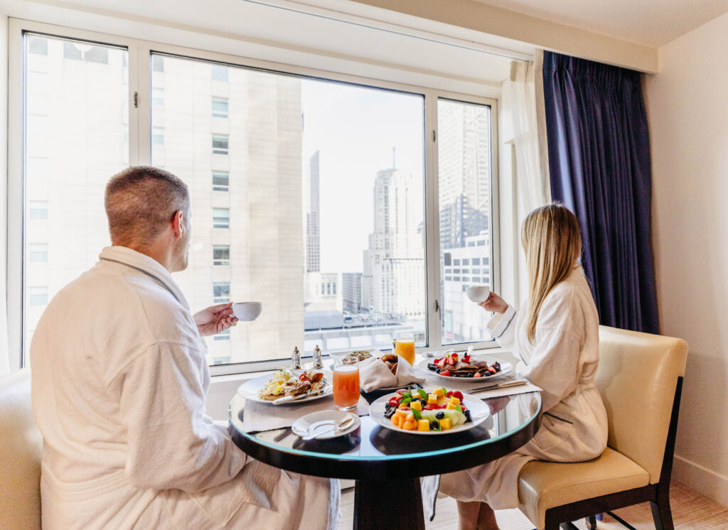 Two people in white robes sit at a round table with breakfast, looking out a large window at city buildings.