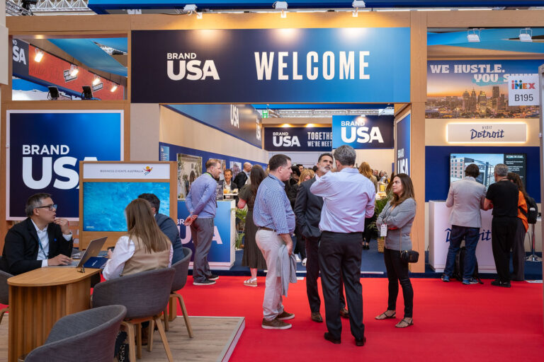 People gather and converse at a “Brand USA” booth in a convention center, with informational displays and signage welcoming visitors.