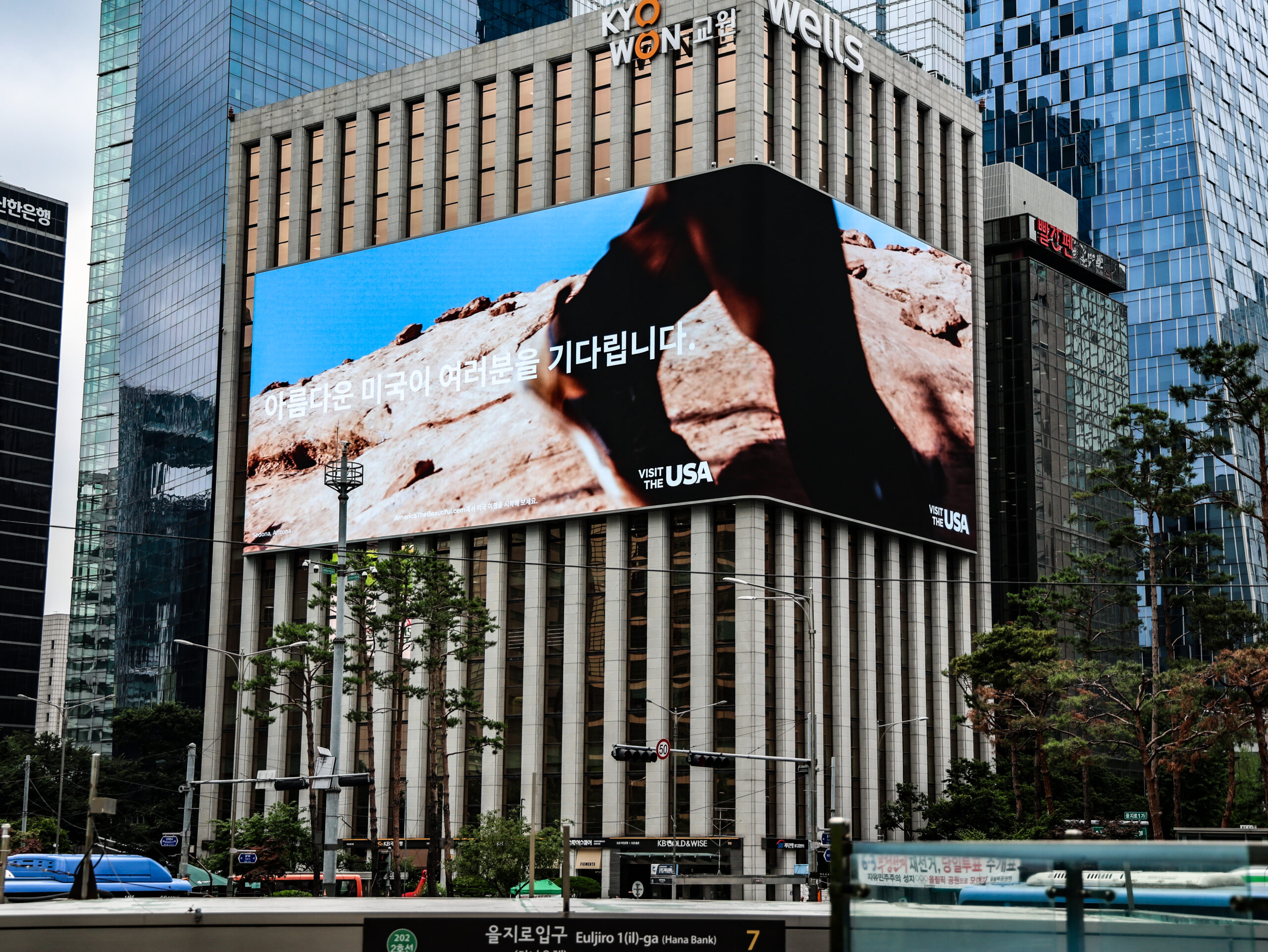 Large digital billboard on a city building displays an ad with a desert scene and Korean text; skyscrapers and trees surround the area.