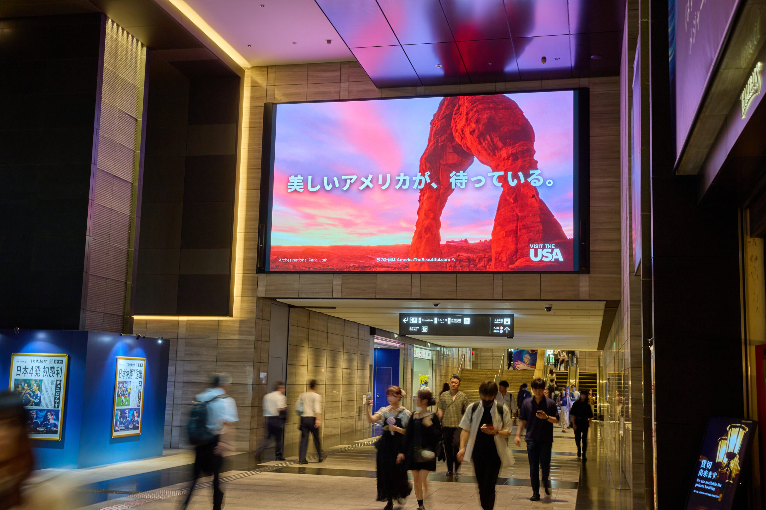 People walk through a modern indoor corridor with a large digital billboard displaying a scenic rock formation and Japanese text.