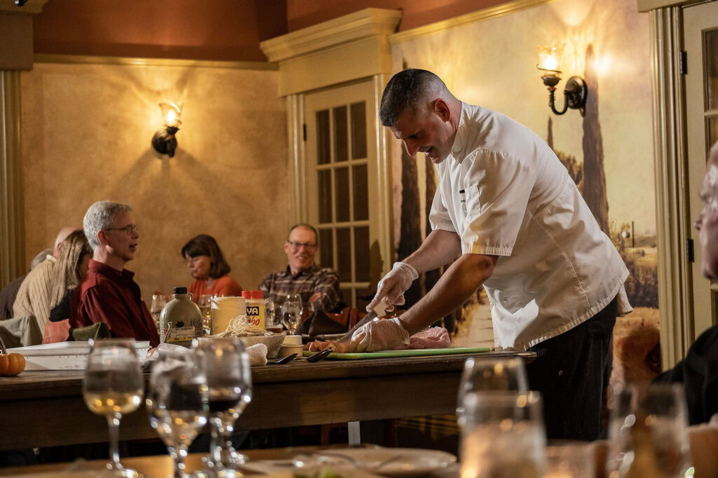 A chef prepares food at a table in front of seated diners in a warmly lit restaurant with wine glasses and tableware visible.