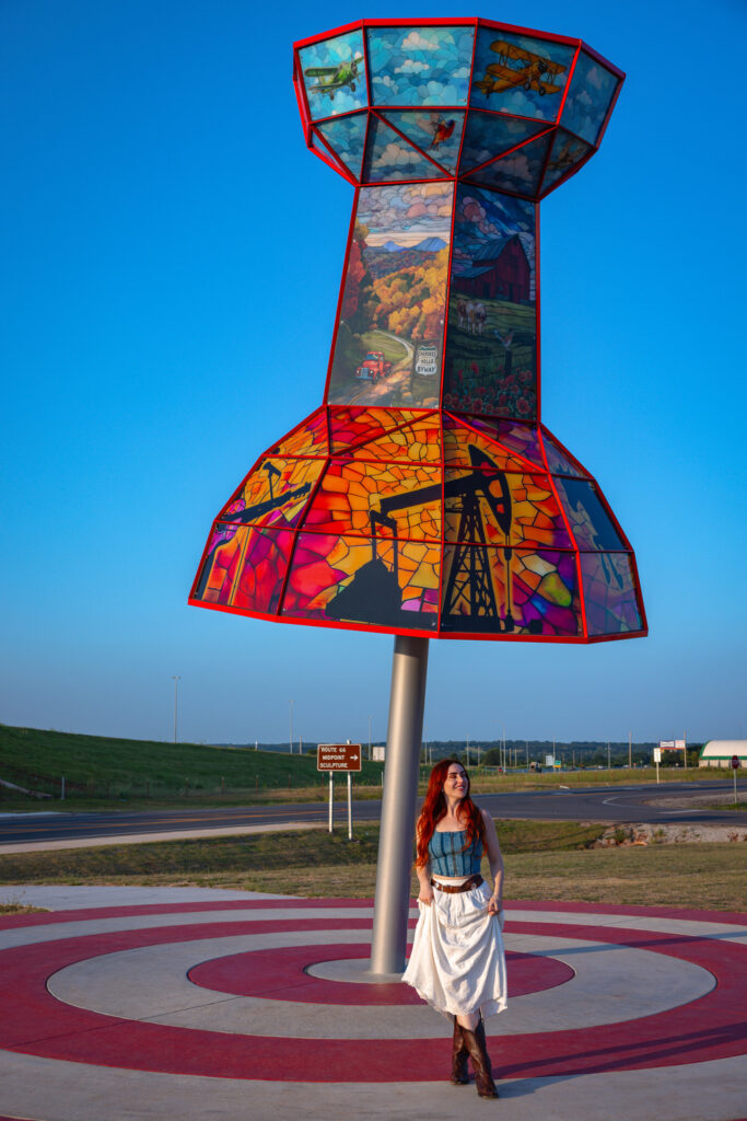 A woman stands in front of a large, colorful pushpin sculpture featuring artwork panels, set outdoors near a road under a clear blue sky.
