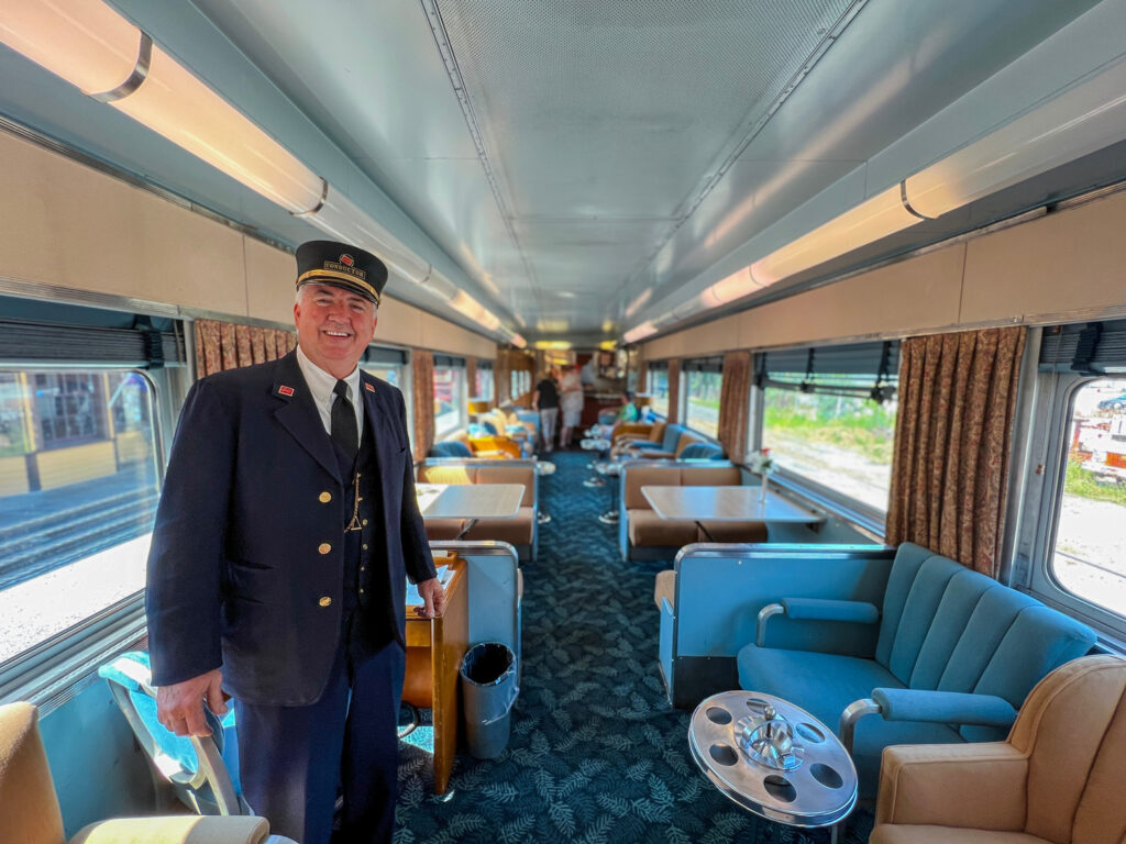 A train conductor in uniform stands inside a vintage passenger car with blue and beige seating, tables, and large windows.