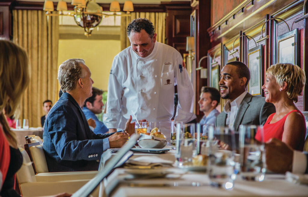 A chef speaks with seated diners at a restaurant table, with others dining in the background. Glasses, plates, and menus are visible on the table.