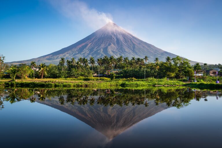 A conical volcano emits a small plume of smoke, reflected in a calm body of water, with palm trees and houses at its base under a clear blue sky.