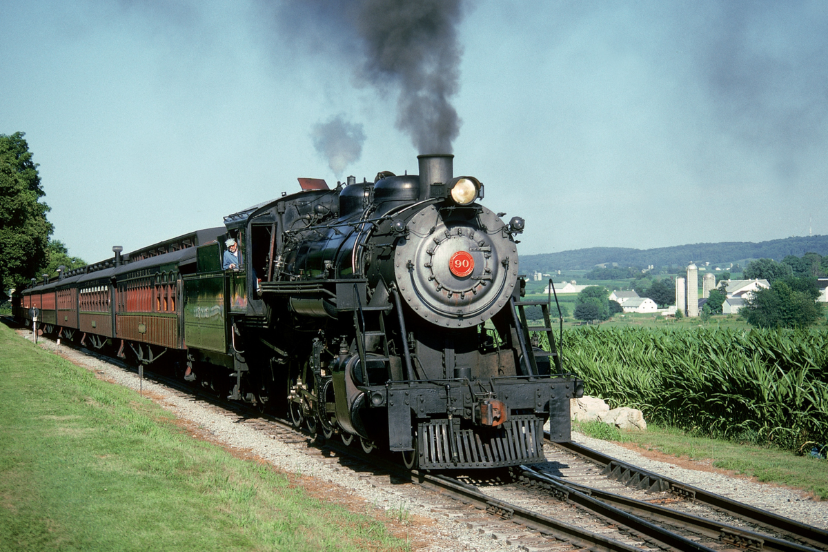A black steam locomotive pulls several passenger cars down a rural track, with fields, trees, and farm buildings in the background under a clear blue sky.