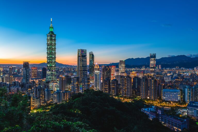 Taipei city skyline at sunset with Taipei 101 and other skyscrapers illuminated, mountains in the background, and trees in the foreground.