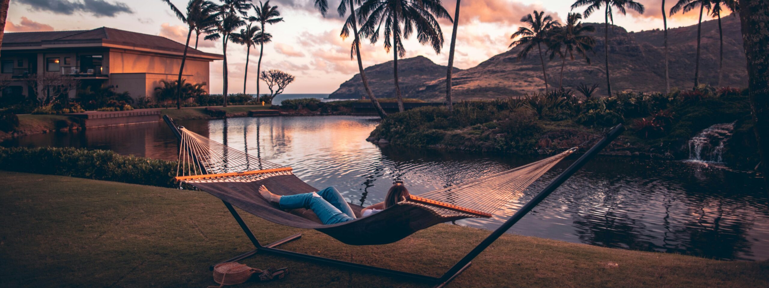 Person relaxing on a hammock by a pond at sunset, surrounded by palm trees, with mountains and a building in the background.
