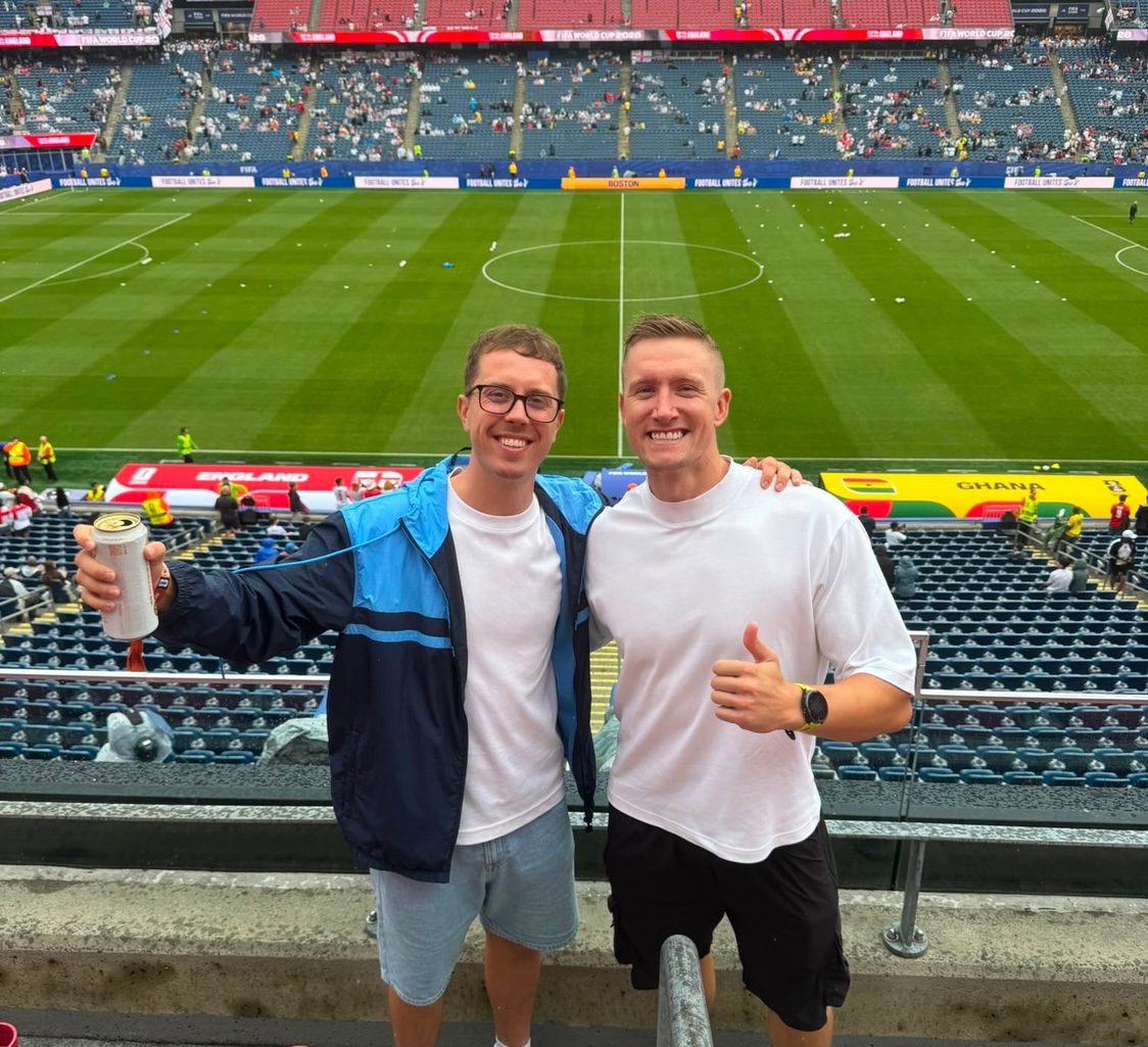 Two men stand smiling in front of a soccer field, with one holding a drink and the other giving a thumbs-up. Crowds fill the stadium seats in the background.