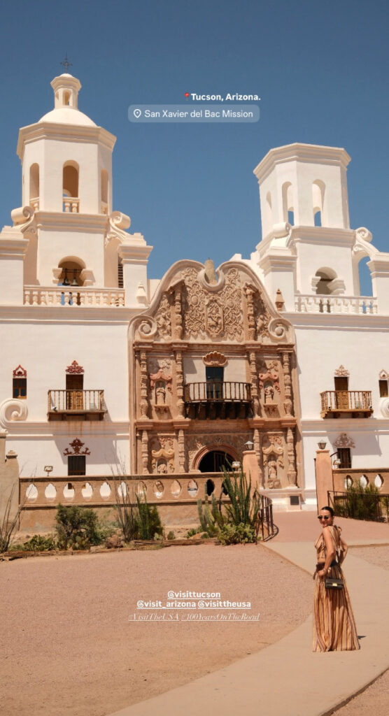 San Xavier del Bac Mission in Tucson, Arizona, with a woman in a long tan dress posing in front of the ornate church facade on a sunny day.