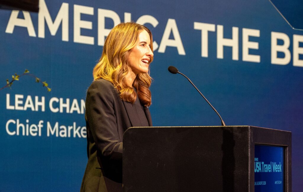A woman stands at a podium speaking into a microphone at an event, with a blue backdrop displaying partial text and her name in the background.