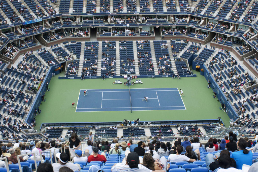 Aerial view of a tennis match in a large stadium with spectators seated throughout the stands and players on a blue and green court.