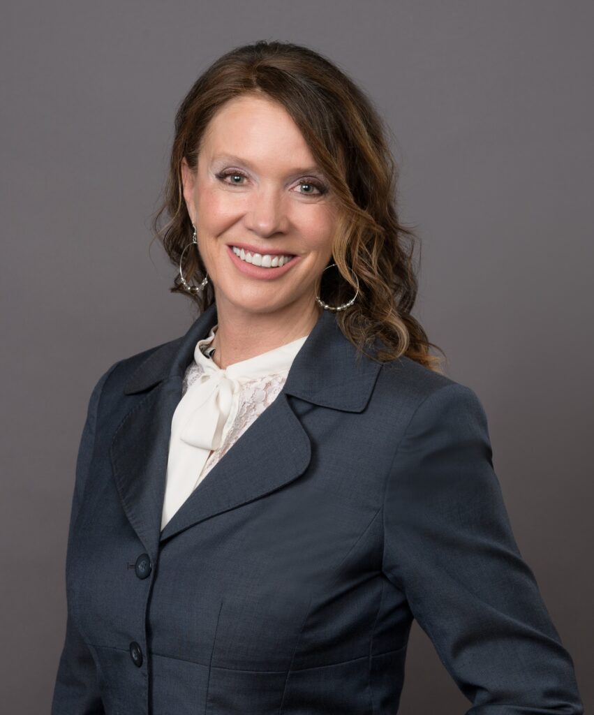 A smiling woman wearing a dark blazer over a white blouse with a tie-neck detail.