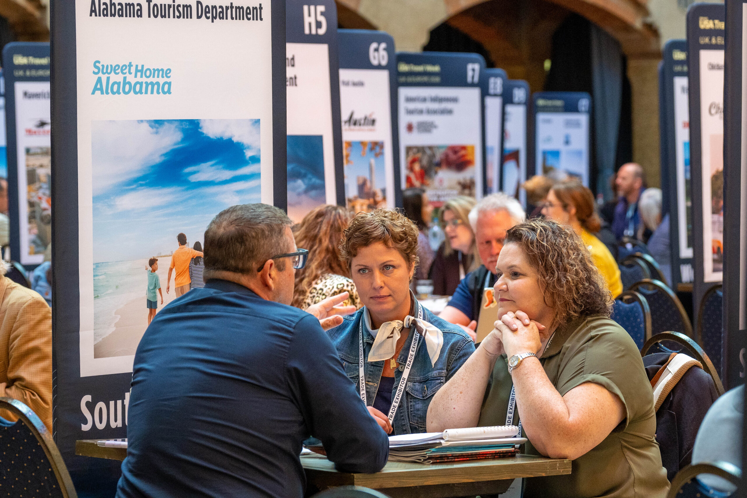 Three people sitting at a table at a trade show, having a conversation in front of booths for the Alabama Tourism Department.