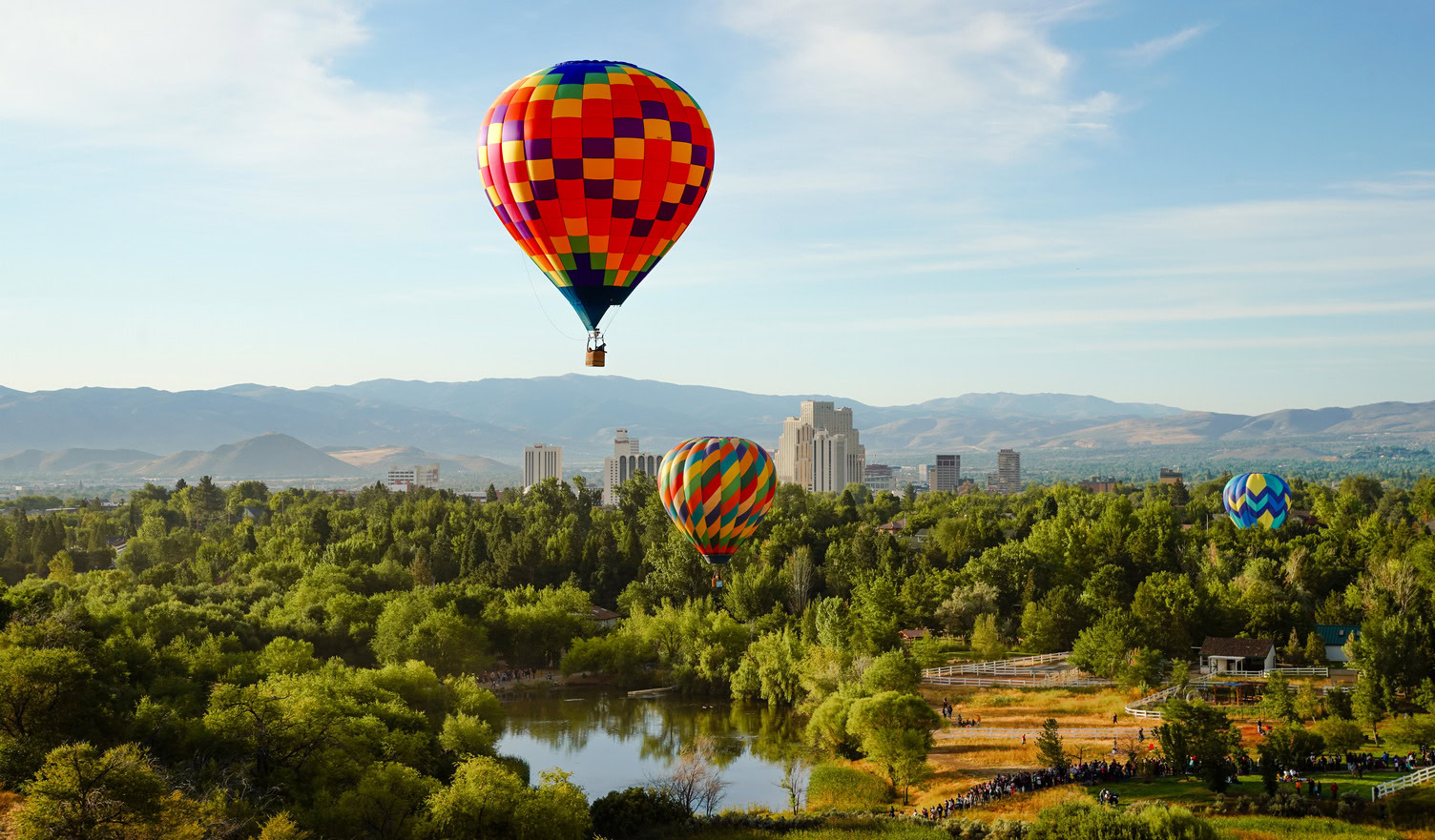 Three colorful hot air balloons floating above a park with a pond and a city skyline in the distance.