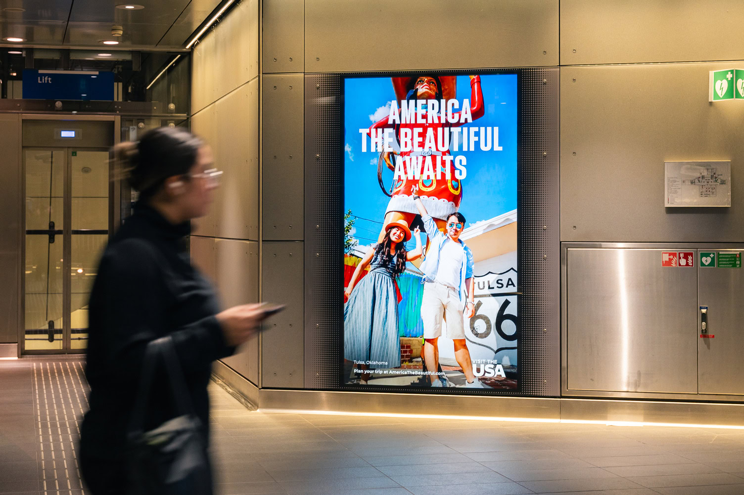A person walks past a digital billboard showing a travel advertisement for Tulsa, Oklahoma, featuring a couple posing in front of a giant statue.