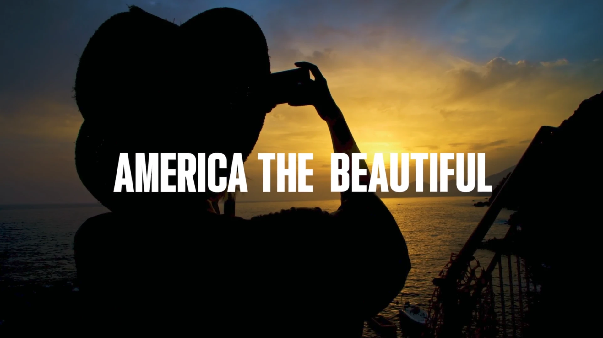 Silhouette of a person wearing a hat taking a photo of a sunset over the ocean, with the text “AMERICA THE BEAUTIFUL” centered.